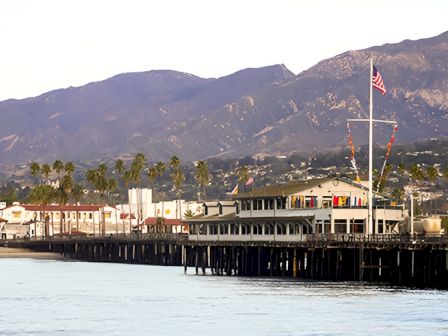 A seaside pier with a long row of buildings, palm trees, mountains in the background, and an American flag atop a flagpole, over calm water.