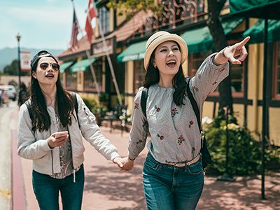 Two women walk hand-in-hand down a sunny street; one points ahead with excitement while the other laughs behind sunglasses.