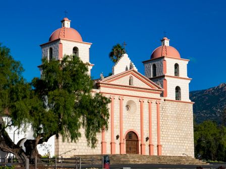 A peach-colored church with two square bell towers, red domes, stone facade, set against a bright blue sky and surrounding trees.