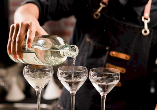 A person pours clear liquid from a bottle into three elegant stemmed glasses, likely a cocktail setup, with a focus on the action.