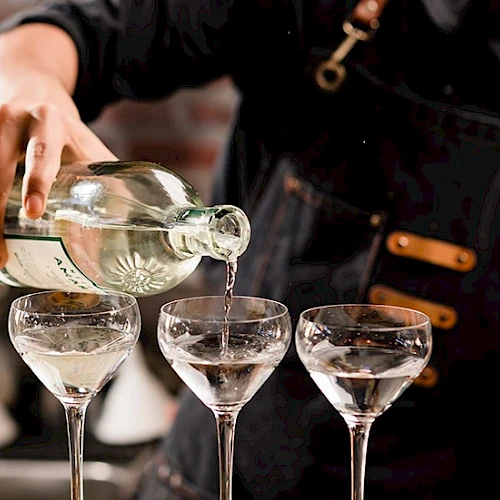 A person pours clear liquid from a bottle into three elegant stemmed glasses, likely a cocktail setup, with a focus on the action.