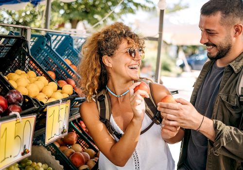 A couple is shopping at an outdoor fruit stand, smiling and inspecting peaches together under a blue canopy.