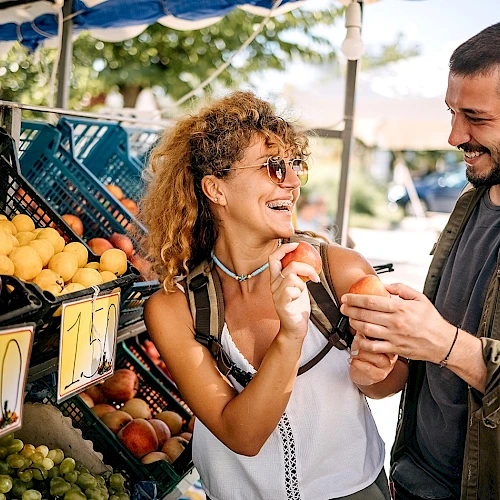 A couple is shopping at an outdoor fruit stand, smiling and inspecting peaches together under a blue canopy.