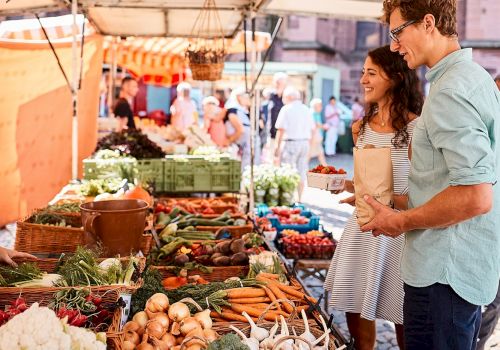 A couple shops for fresh vegetables at an outdoor market, browsing colorful produce like carrots, greens, and onions under a stall canopy.