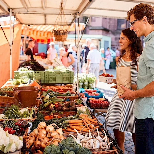 A couple shops for fresh vegetables at an outdoor market, browsing colorful produce like carrots, greens, and onions under a stall canopy.