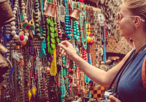Colorful bead necklaces and trinkets on a market stall; a shopper examines them closely, jewelry galore in vibrant hues.
