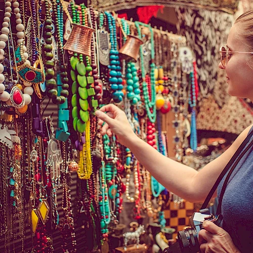 Colorful bead necklaces and trinkets on a market stall; a shopper examines them closely, jewelry galore in vibrant hues.
