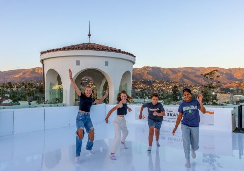 Four friends skate on an outdoor rink with a small round building and mountains in the background, smiling and posing happily.