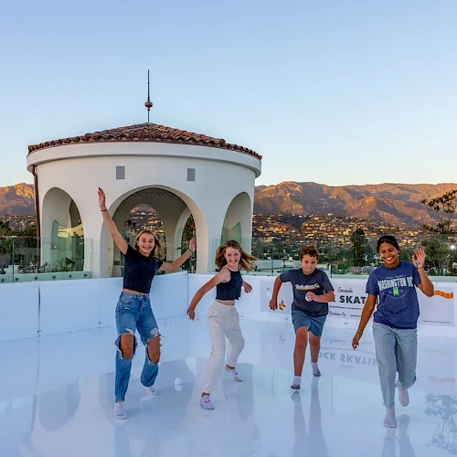 Four friends skate on an outdoor rink with a small round building and mountains in the background, smiling and posing happily.
