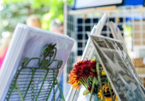 A display stand with magazines and newspapers, featuring flowers in a sunny outdoor setting, likely at a market or cafe.