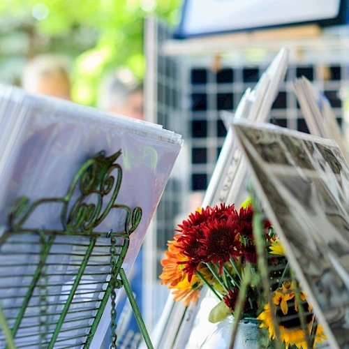 A display stand with magazines and newspapers, featuring flowers in a sunny outdoor setting, likely at a market or cafe.