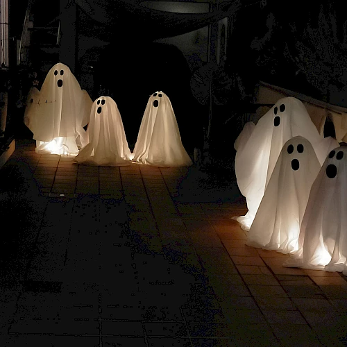 Silhouetted ghostly figures drift along a dark path, illuminated by warm lanterns, creating a spooky, seaside funeral-like procession.