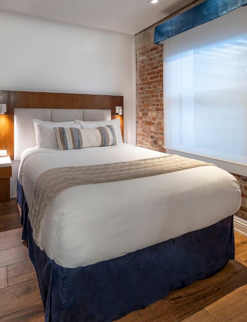 A tidy hotel room with a queen bed, exposed brick wall, wooden floor, desk and chair, wall-mounted TV, and a bright window beside the bed.