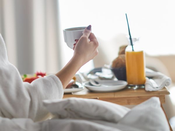 A person in bed holding a teacup with a breakfast tray: orange juice, fruit, toast, and a striped straw against a bright, cozy morning backdrop.
