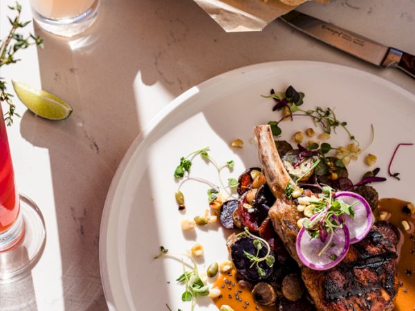 A plated dish with grilled meat, onions, herbs, a tomato-orange sauce, and tortilla chips nearby; lime wedge and drinks on a bright table.
