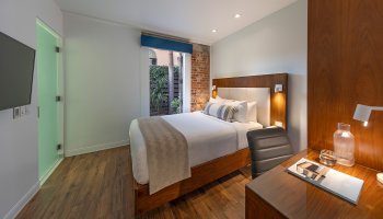 A modern hotel bedroom with a wooden bed, white linens, a nightstand lamp, a glass door to the left, and a desk with a chair, all on wood flooring.