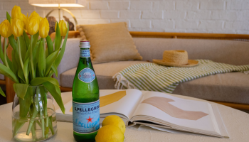A cozy living room scene: a vase of yellow tulips, a bottle of San Pellegrino, a lemon, a magazine open on a coffee table, and a striped throw on the sofa.