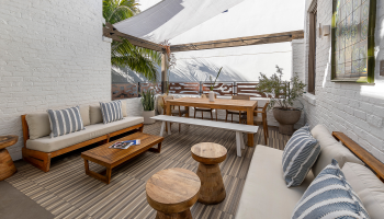A bright outdoor patio with white brick walls, wooden furniture, a long dining table, cushioned seating, striped pillows, and potted plants under a shade sail. (140 chars)