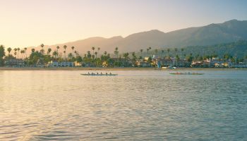 A calm coastal scene at sunset with palm trees, boats on the water, and distant mountains lining the shore.