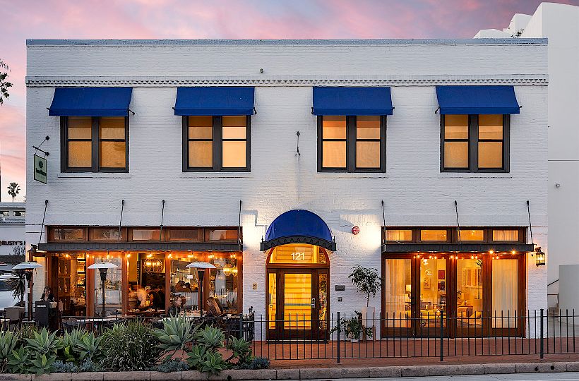 A charming white storefront with blue awnings, warm-lit windows, and outdoor seating; palm trees frame the scene at dusk.