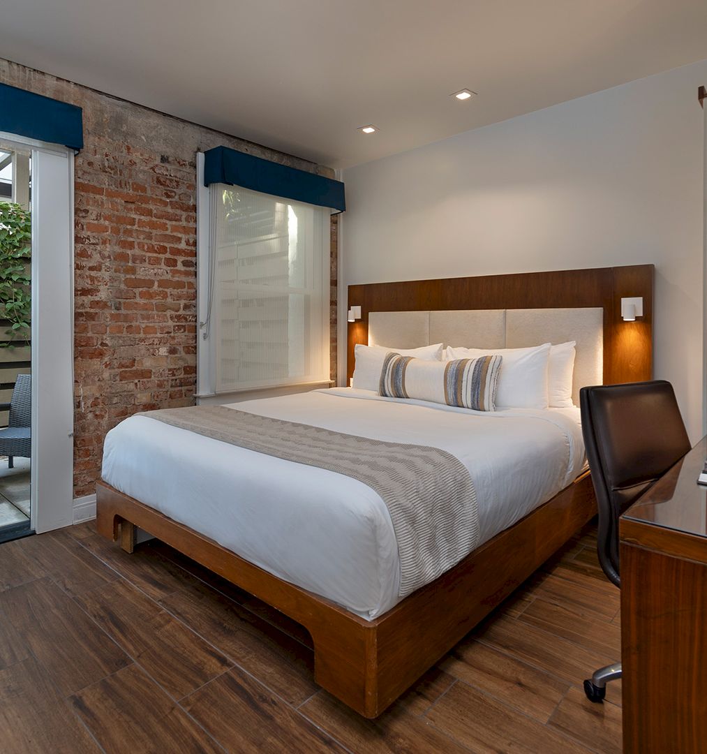 A modern hotel room with a large bed, wooden furniture, a work desk, and a small outdoor patio area visible through glass doors.