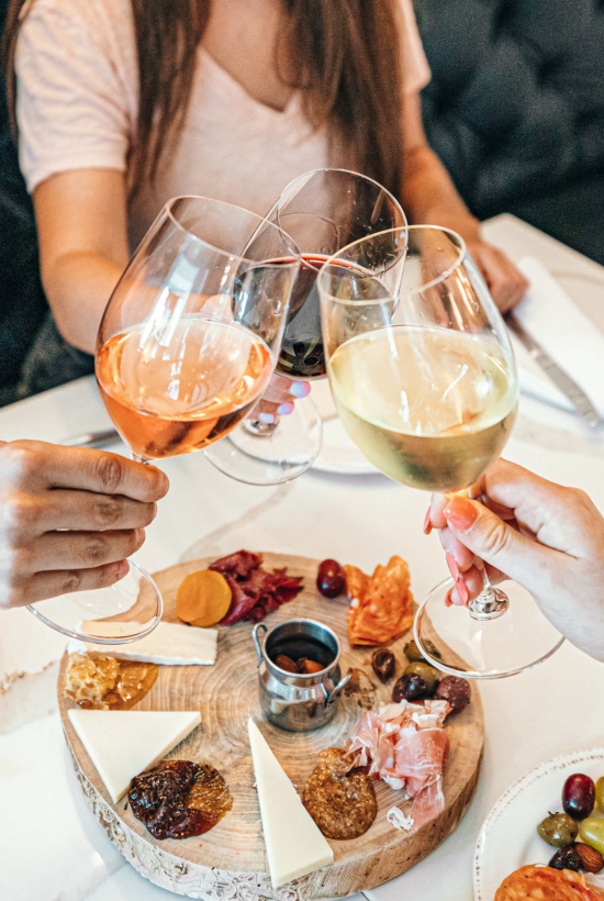 People toast wine glasses around a cheese board with assorted bites on a round wooden platter at a cozy table.