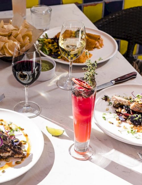 A fancy table with multiple plated dishes, wine glasses, a bright red cocktail, chips, and a sunny, upscale dining setting.