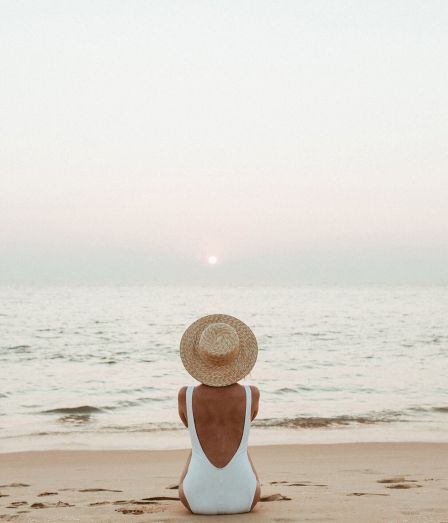 A person in a white swimsuit sits on the sandy beach, facing the calm sea at sunset, wearing a straw hat as the sun dips over the horizon.
