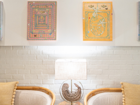 A cozy living room with a white brick wall, four framed colorful geometric artworks, a lit table lamp, and two beige sofas with pom-pom pillows facing each other.