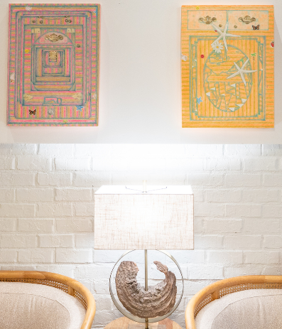 A cozy living room with a white brick wall, four framed colorful geometric artworks, a lit table lamp, and two beige sofas with pom-pom pillows facing each other.