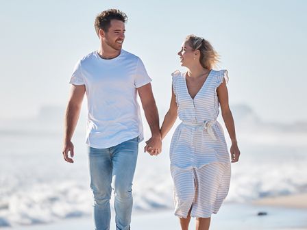 A couple walking hand in hand on the beach, smiling and enjoying a sunny day by the ocean.