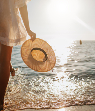 A person stands at the shore holding a wide-brim straw hat, waves gently lap the beach, sun setting over the calm sea.
