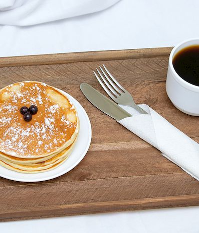 A wooden tray with a stack of pancakes topped with berries, a cup of black coffee, a fork, and a napkin beside them, ready to enjoy.