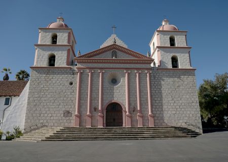 A stone church with pale pink trim, two square bell towers, a wide staircase, and a triangular pediment under a blue sky.
