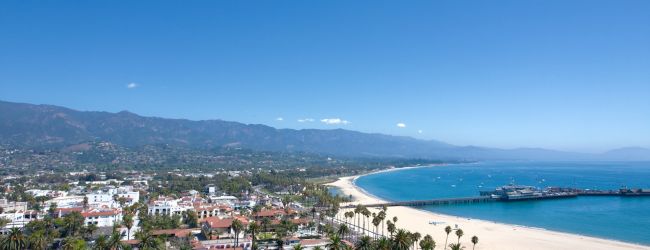 A sunny coastal town with white buildings, a sandy beach, palm trees, and blue ocean under a clear sky.