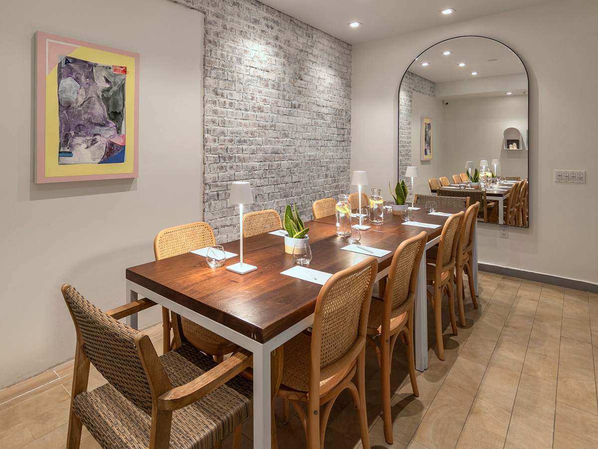 A modern dining nook with a long wooden table and wicker chairs, soft lighting, brick accent wall, framed art, and a tall mirror reflecting the room.