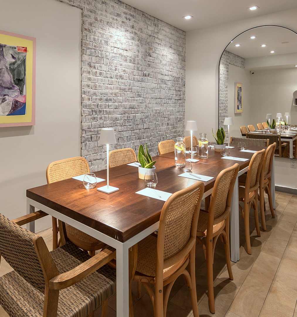 A modern dining nook with a long wooden table and wicker chairs, soft lighting, brick accent wall, framed art, and a tall mirror reflecting the room.