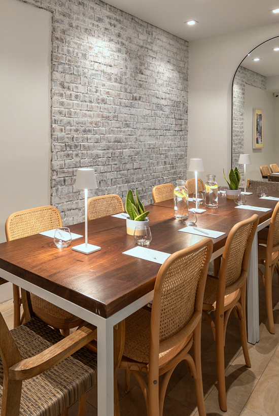 A modern dining nook with a long wooden table and wicker chairs, soft lighting, brick accent wall, framed art, and a tall mirror reflecting the room.