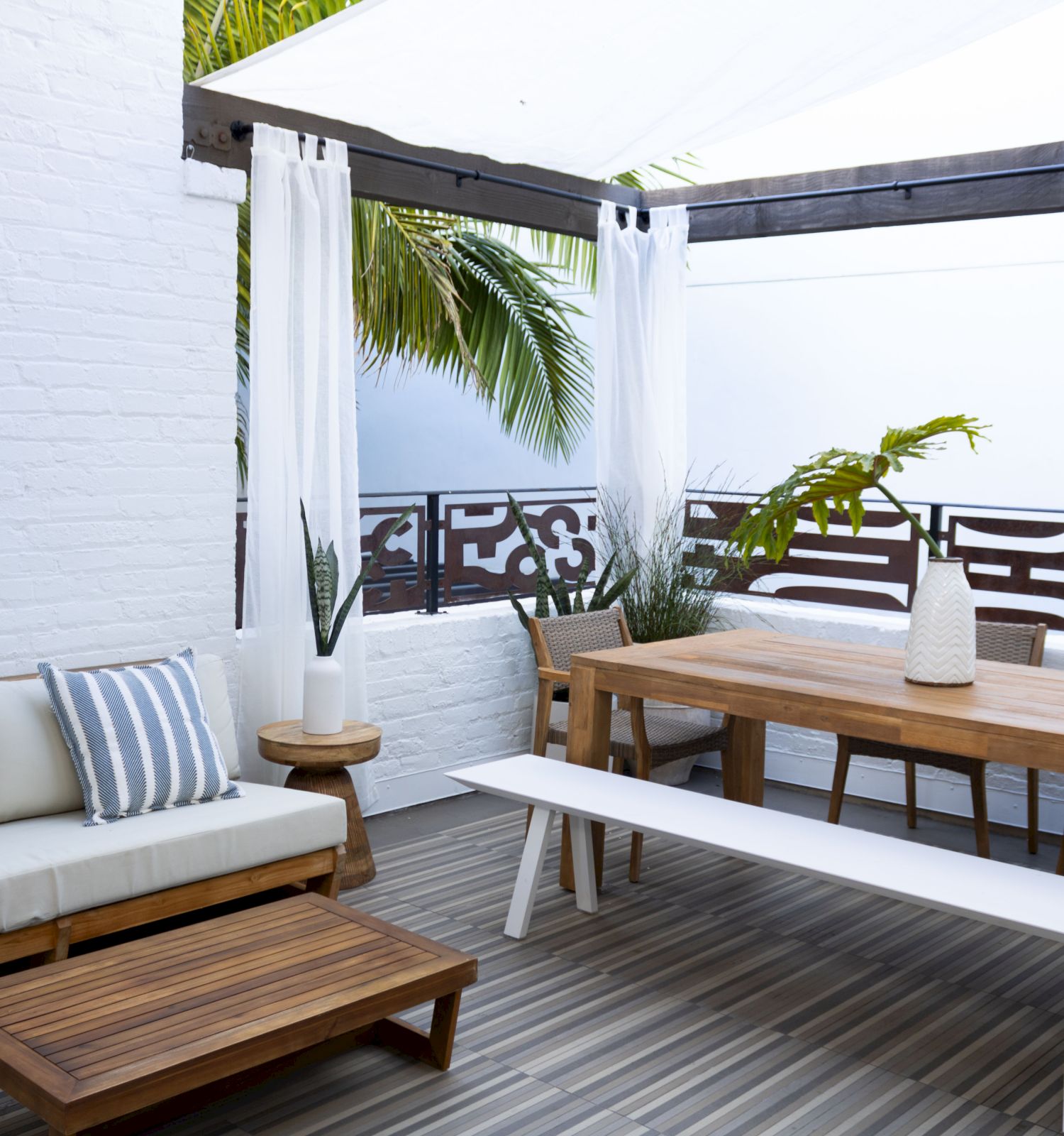 A stylish outdoor patio with a wooden deck, white walls, a beige couch with striped cushions, a long wooden table, white bench, and a shaded canopy with tropical plants.