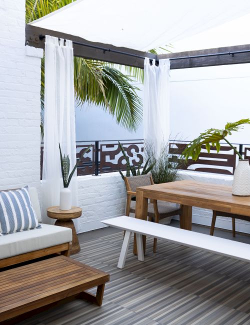 A stylish outdoor patio with a wooden deck, white walls, a beige couch with striped cushions, a long wooden table, white bench, and a shaded canopy with tropical plants.