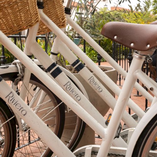 A row of white bicycles with brown saddles and wicker baskets, labeled &ldquo;Rivelo,&rdquo; parked closely together.
