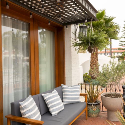 Cozy outdoor seating on a balcony: a wooden-framed sofa with gray cushions and striped pillows, potted plants, and a covered trellis above.