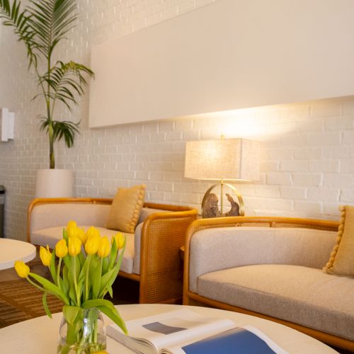 A bright, modern living room with wicker furniture, yellow-tinted cushions, a white brick wall, a potted palm, a lamp, a vase of yellow tulips, and an open magazine on a coffee table.