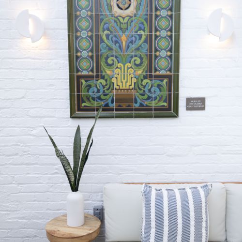 A stylish corner with a framed stained-glass art piece, white brick wall, two wall lights, a small wooden side table with a plant, and a white sofa with blue striped cushions.