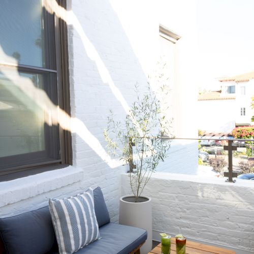 An outdoor balcony with a blue cushioned sofa, striped pillow, a small wooden table, potted plant, and a plate of fruits on a sunny day, ending with a period.