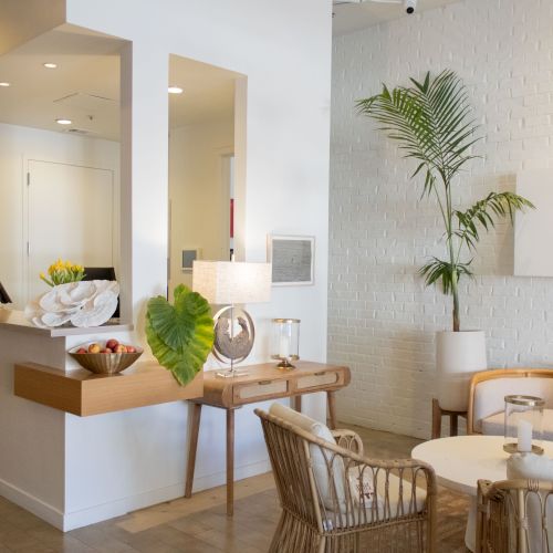 A modern, bright hotel lobby with a white reception desk, a seating area featuring a round table, wicker chairs, a sofa, and green plants, creating a airy, welcoming vibe.