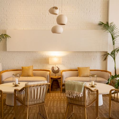 A bright, cozy lounge with wicker chairs, round white tables, potted palms, warm lighting, and soft yellow cushions.