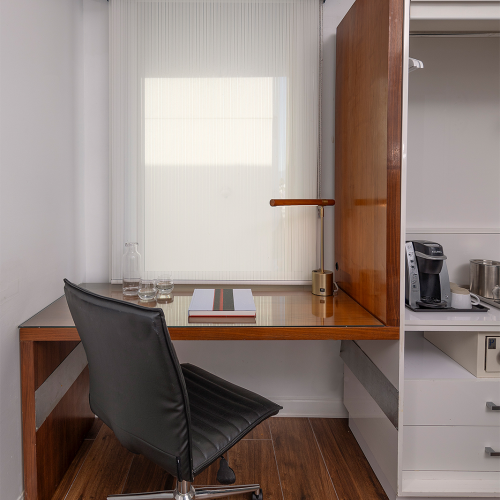A compact office nook with a wooden L-shaped desk, black swivel chair, glass, lamp, and a tidy adjacent kitchenette area behind a partial wall.