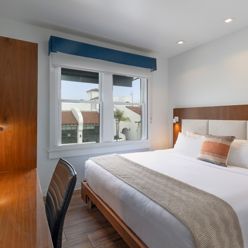 A modern hotel room with a large bed, wooden headboard, side lamps, a desk with a chair, a TV on the wall, and a window with a blue valance.
