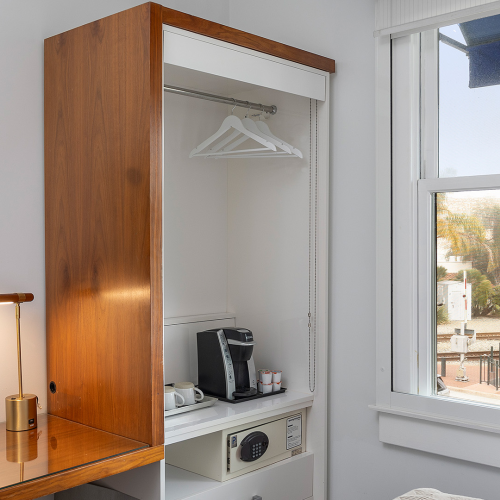 A hotel-style room with a mounted TV, a wooden open wardrobe, a coffee machine inside, a safe, and a desk lamp by a window overlooking palm trees.